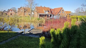Eine Reihe traditioneller Backsteinhäuser mit roten Ziegeldächern, ein kleiner Teich und üppiges Grün im Vordergrund.