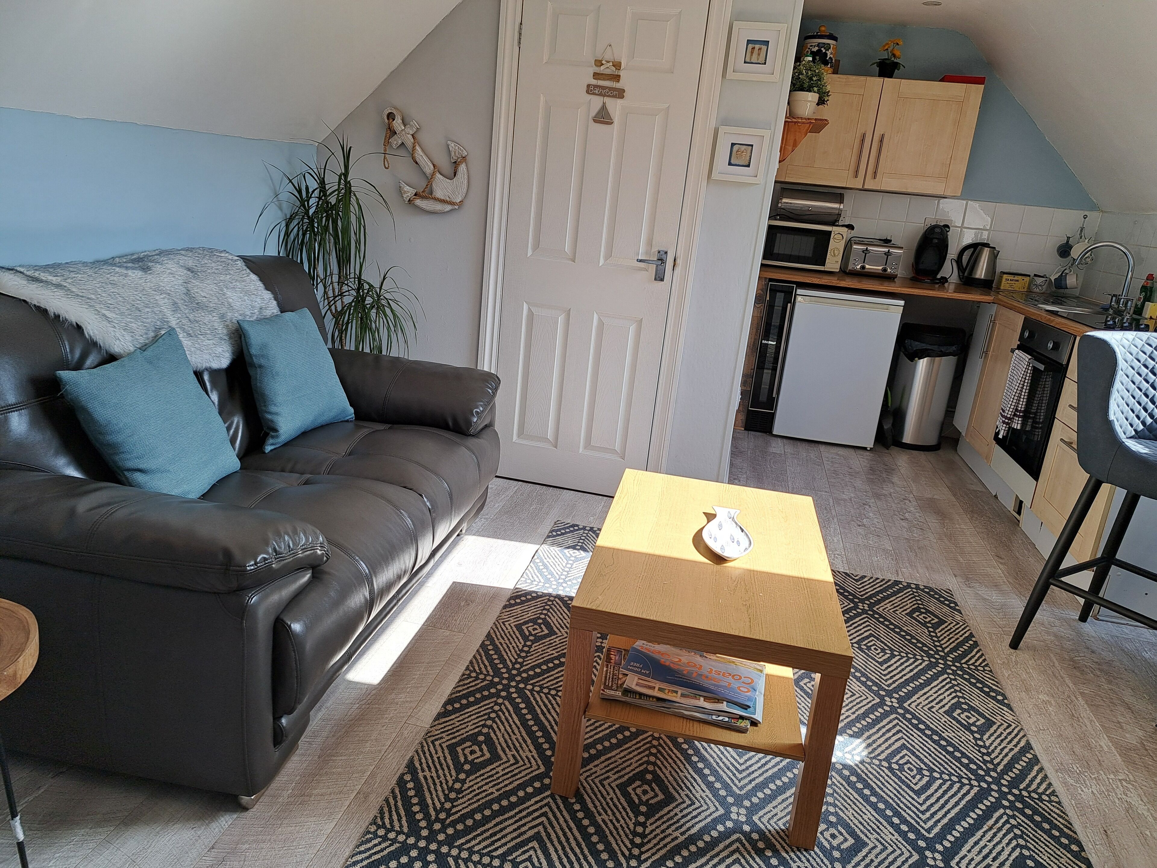 A living room with a dark leather sofa, a small wooden coffee table, and a patterned rug. A kitchen is visible in the background with wooden cabinets and a microwave.