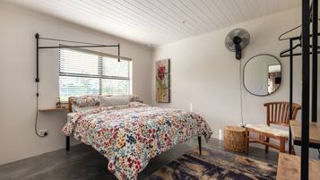 A bedroom with a bed, a standing fan, a chair, and a small table.