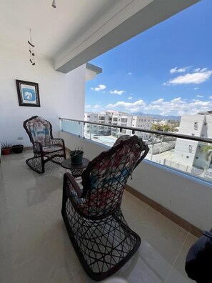 A balcony with two wicker chairs, a small table, and potted plants. - Bello Apartamento en Santiago con Todas las Comodidades (Santiago de los Caballeros)