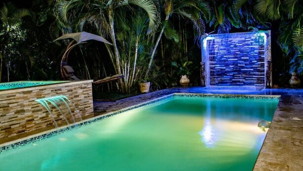 A swimming pool with a waterfall feature, surrounded by palm trees and a stone wall. - Casa Linda Beautiful Villa (Sosúa)