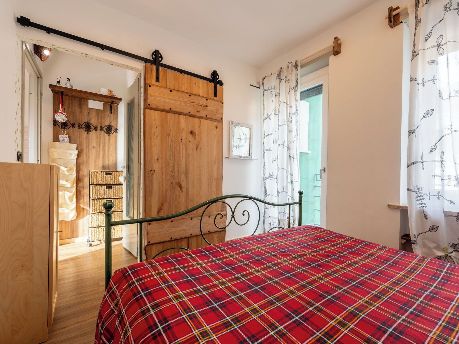A bedroom with a plaid bedspread, a wooden wardrobe, a metal bed frame, and a view of the outside through a door.