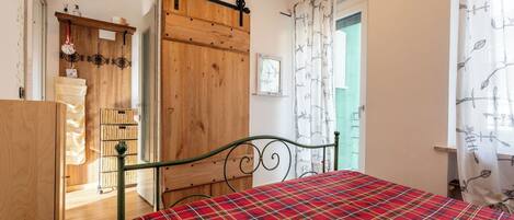 A bedroom with a plaid bedspread, a wooden wardrobe, a metal bed frame, and a view of the outside through a door.