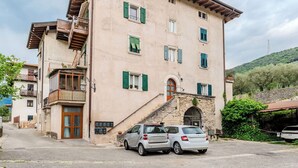 Un immeuble à plusieurs étages, avec un escalier en pierre menant à l’entrée, une voiture garée devant et une colline avec des arbres en arrière-plan.