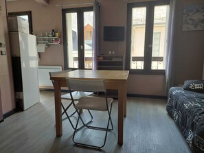 Une petite pièce bien éclairée, avec une table en bois et une seule chaise, un canapé agrémenté de coussins à motifs, un téléviseur fixé au mur et un réfrigérateur dans un coin.