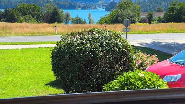 A view from a covered porch overlooking a lake, mountains, and a red car parked nearby. - Holiday Home LiRa (Villach)