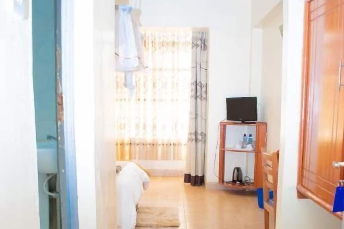 Un pasillo con suelo de baldosas, un armario de madera, un televisor sobre un mueble y una cama con una colcha blanca.