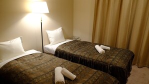 Ein Hotelzimmer mit zwei Betten, jedes mit einer gemusterten Tagesdecke und weißen Kissen. Auf der linken Seite des Zimmers steht eine Lampe, auf der rechten eine Gardine.