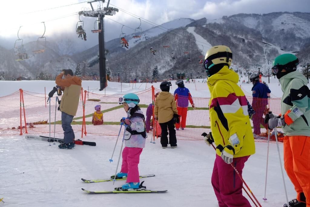 A ski slope with people skiing and standing, a ski lift in the background, and a mountain range.