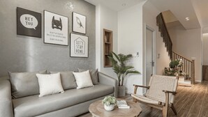 Un salon moderne avec un canapé gris, une table basse en bois et un fauteuil en osier. Des tableaux encadrés ornent le mur, et un escalier se trouve au fond de la pièce.
