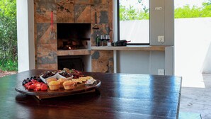 A kitchen with a stone fireplace, a dining table with a fruit platter, and a window with a view of greenery.