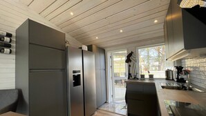 A modern kitchen with a built-in stainless steel refrigerator, a wooden dining table, and a view of the outdoors through a large window. - 5 Star Holiday Home in Skummeslovsstrand (Skummesloevsstrand)