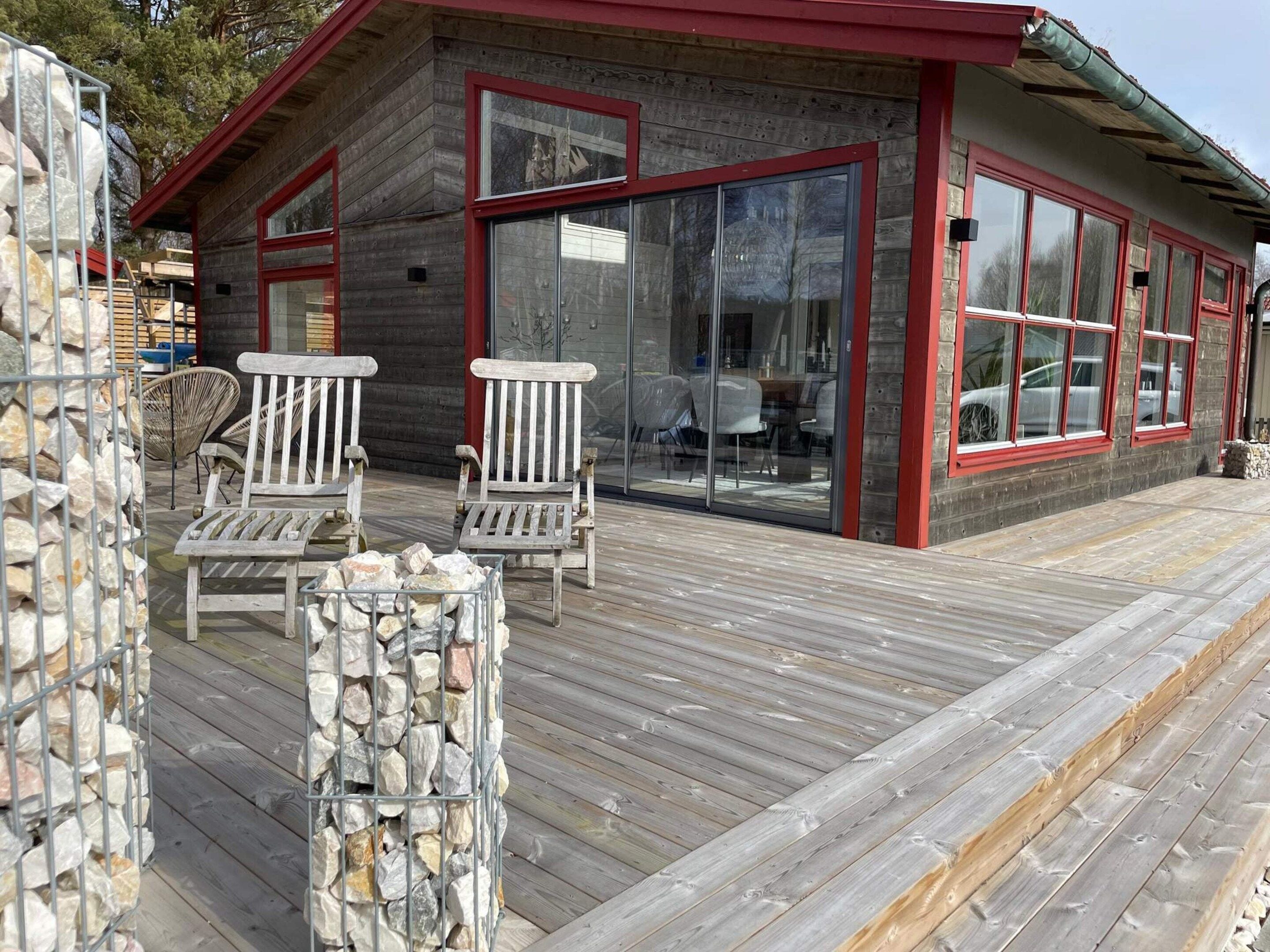 A wooden cabin with a deck, two white chairs, and a stone wall.