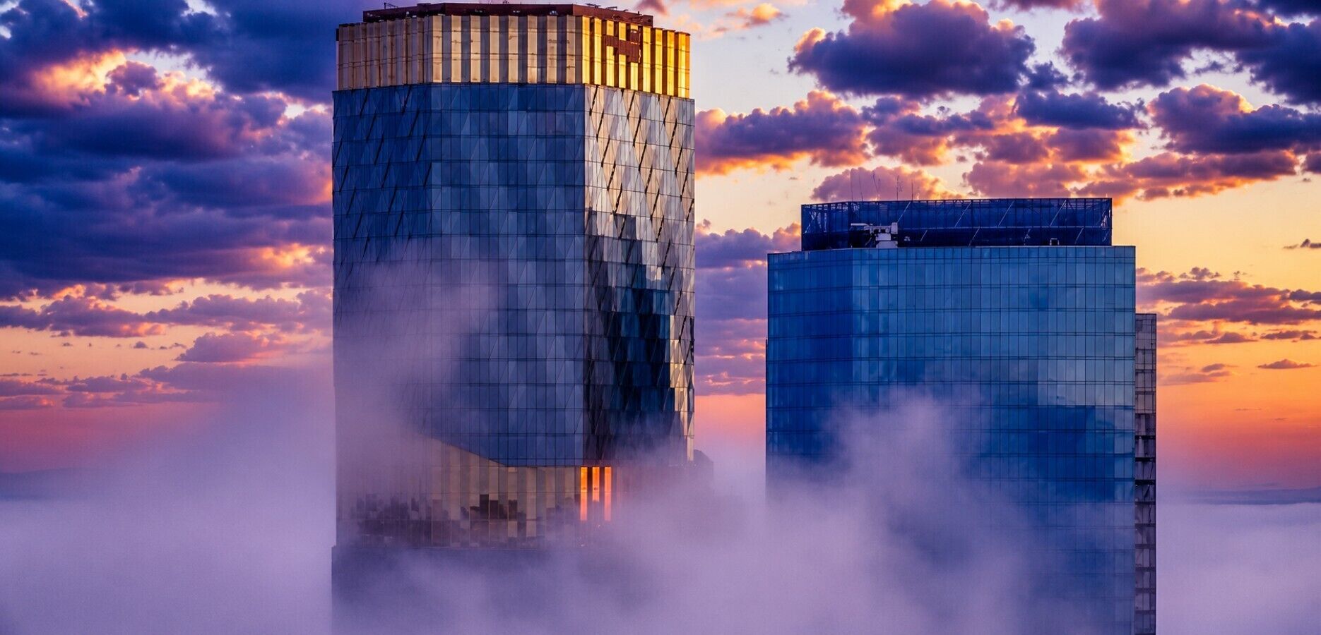 Two high-rise buildings with a reflective glass facade, surrounded by clouds at sunrise or sunset.