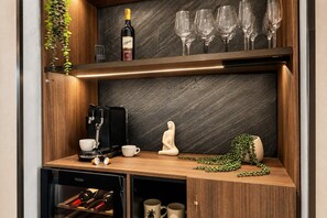 A compact kitchen area with a coffee machine, wine bottle, and glasses on a dark backsplash. - The Skyline Grand | Panoramic Views + Free Parking! (Melbourne)
