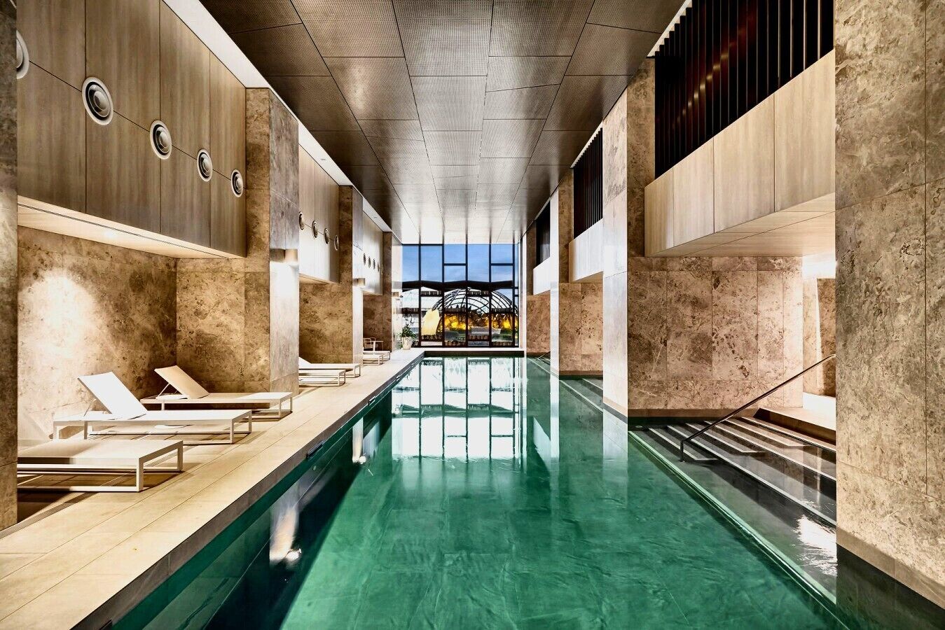 A modern indoor swimming pool with lounge chairs and a view of a sunset through a glass structure.