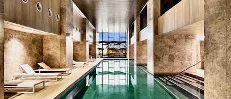 A modern indoor swimming pool with lounge chairs and a view of a sunset through a glass structure.