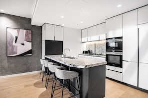 A modern kitchen with a central island, bar stools, stainless steel appliances, and a marble countertop. - The Skyline Grand | Panoramic Views + Free Parking! (Melbourne)