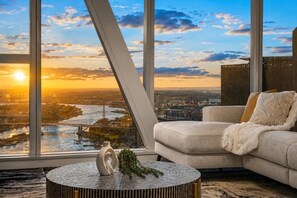 A modern living room with a large window overlooking a cityscape at sunset. - The Skyline Grand | Panoramic Views + Free Parking! (Melbourne)