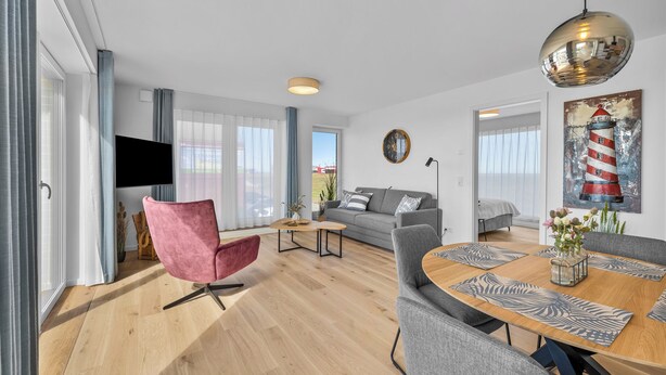 A modern living room with a sofa, a pink chair, a wooden dining table with placemats, a round coffee table, and a large mirror. - The View G01 Licht-Luft-Ebbe und Flut - Balcony, Barbecue, Parking Space (Dagebüll)
