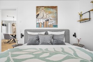 Ein modernes Schlafzimmer mit einem großen Bett, grauen Kissen, einem Wandbild mit Vögeln und einem Nachttisch mit einer Vase.