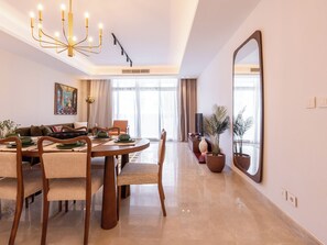 A modern dining area with a round wooden table, cushioned chairs, and a chandelier. There is a large mirror on the wall and a potted plant near the window. - Chic 2BR Apart Garden view Cairo Festival City (Cairo Governorate)