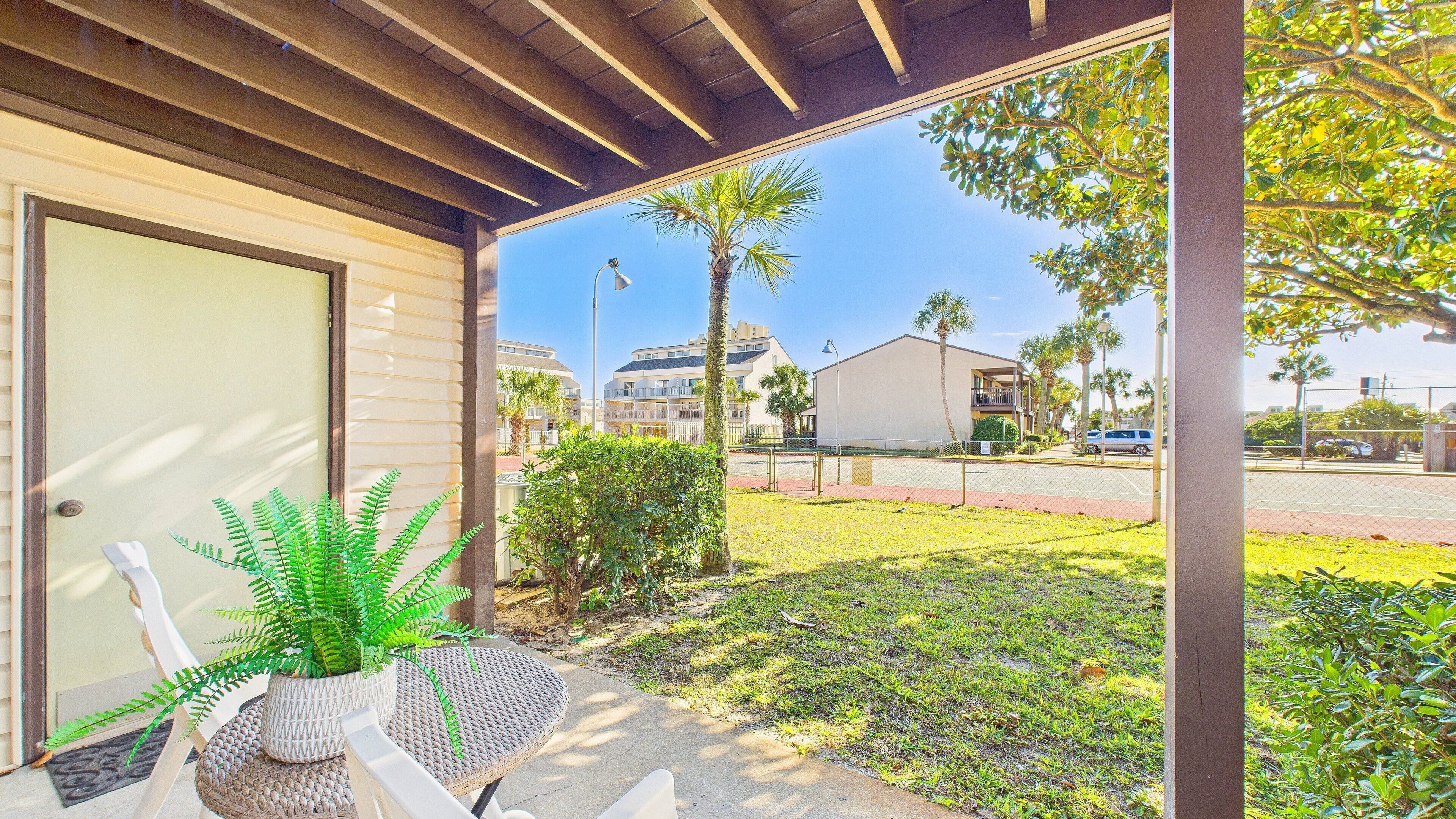 A covered porch with a potted plant, a wicker chair, and a view of a street with buildings and palm trees.