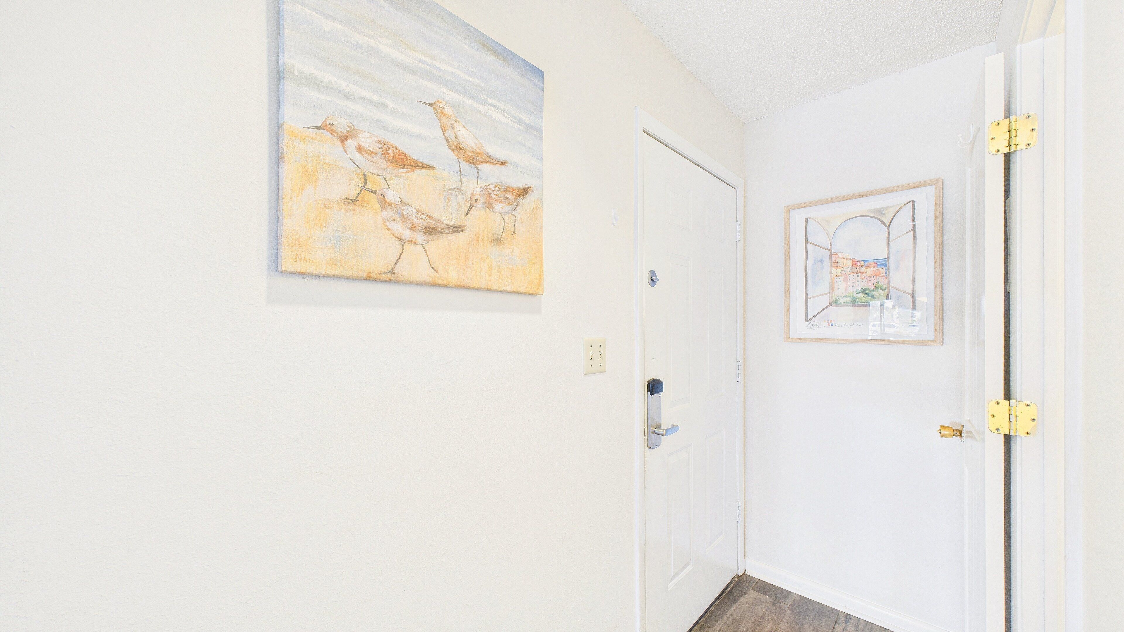 Un couloir avec une porte blanche, un tableau représentant des oiseaux et une photographie encadrée d’un paysage urbain.
