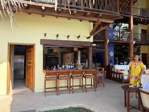 An outdoor bar area with wooden chairs and a bar counter, a balcony with a railing, and a building entrance. - Terrific 4-bedroom villa, private pool and Zihua bay views. Service included! (Zihuatanejo)