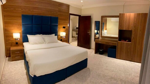 A modern hotel room with a large bed, wooden headboard, bedside lamps, a vanity with a mirror, and a walk-in closet. - Amaken For Residential Units (Al-Hofuf)