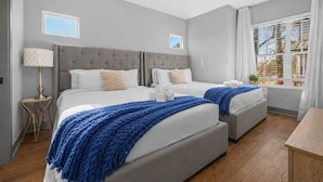 Hotellihuone, jossa on kaksi sänkyä, ikkuna, jossa on sälekaihtimet, yöpöytä lampun kanssa ja pieni pöytä, jossa on kasvi.