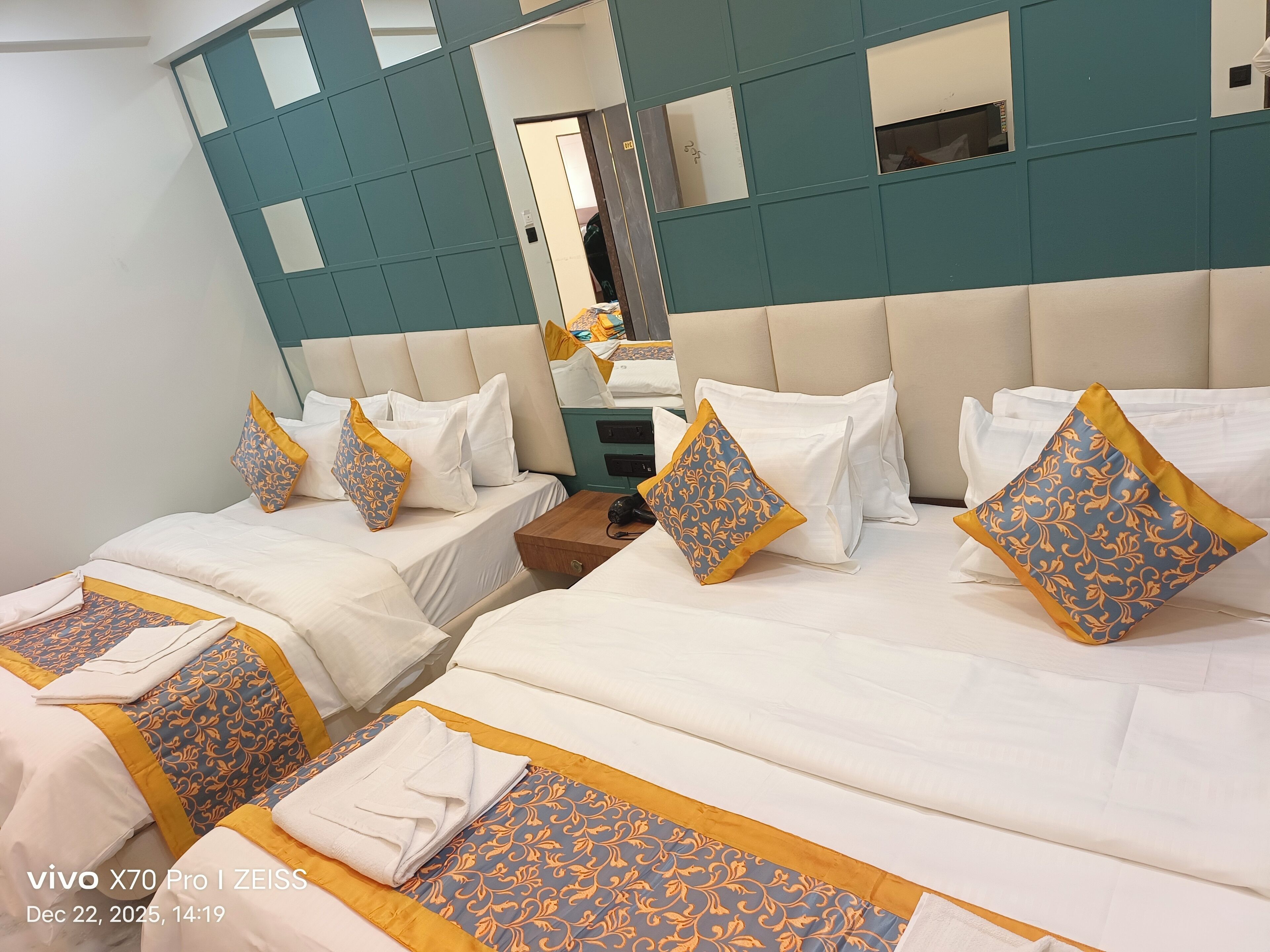A hotel room with two beds, each with white linens and decorative pillows. There is a wooden nightstand with a lamp and a folded towel. The room has teal-colored walls and a large mirror above the beds.