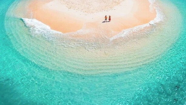 Vista aérea de una pequeña isla circular con una playa arenosa y aguas turquesas transparentes.