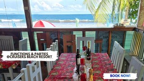 Comedor en Cruisers Beach Resort con vista al océano, mesas dispuestas con copas y botellas de vino, y un paraguas rojo y blanco.