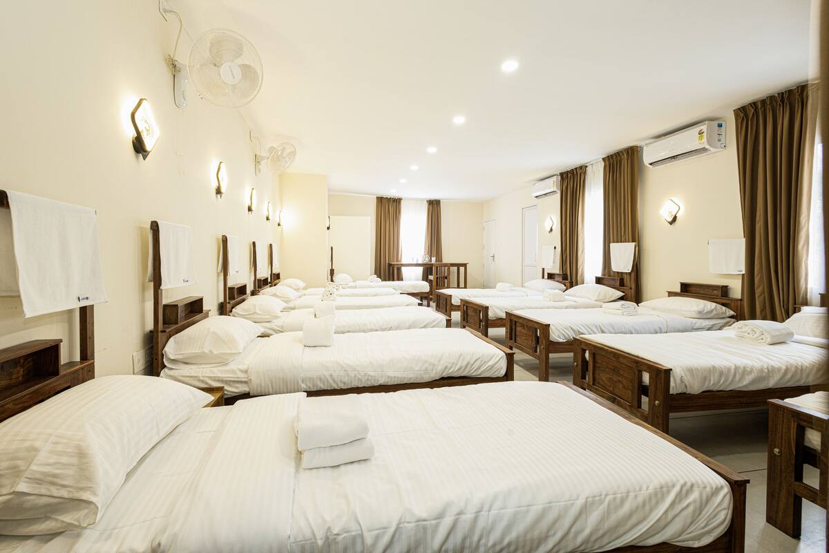 A hotel room with multiple beds, each with white linens and a neatly made towel. There are wall-mounted fans and an air conditioning unit.