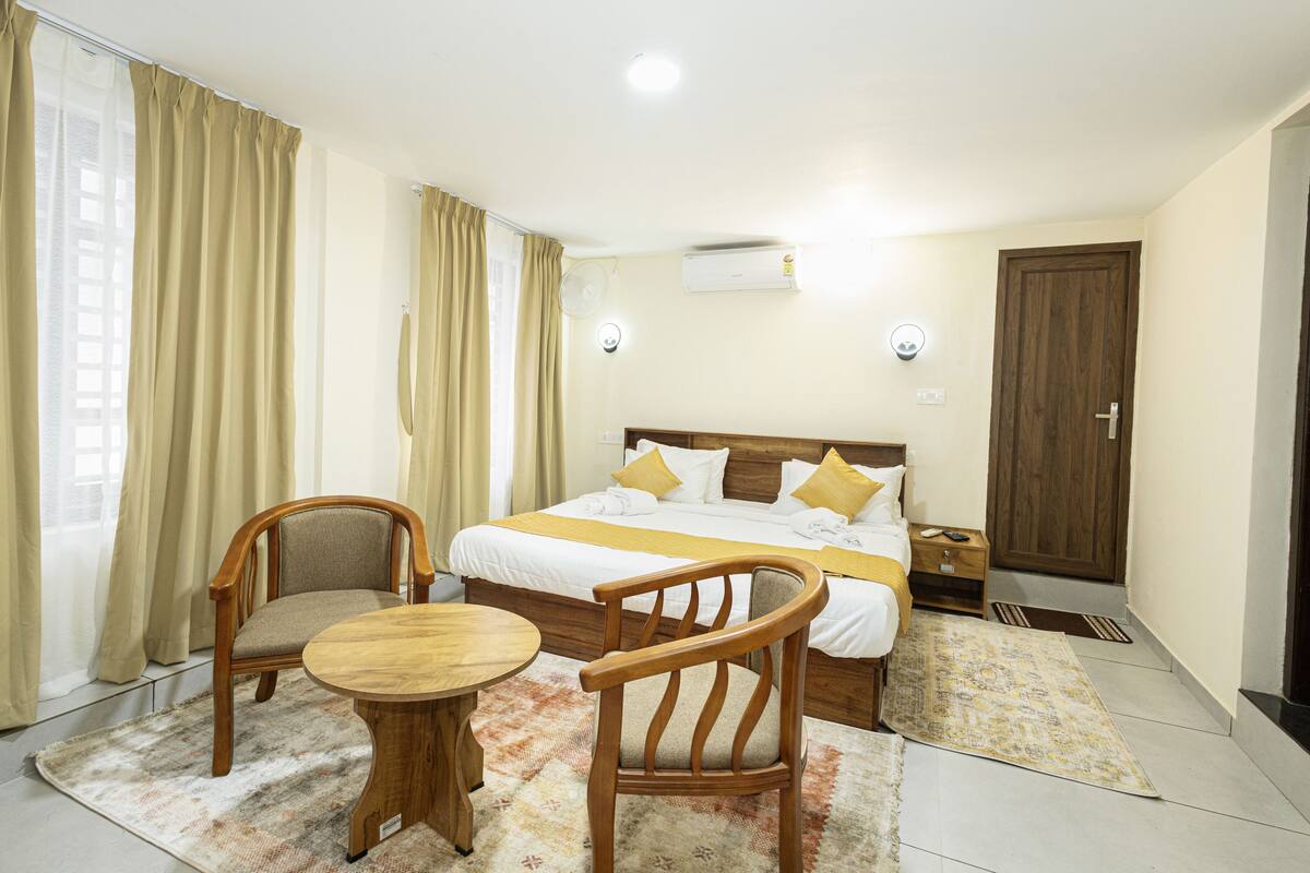 A hotel room with a bed, two wooden chairs, a small round table, a window with curtains, an air conditioning unit, and a door.