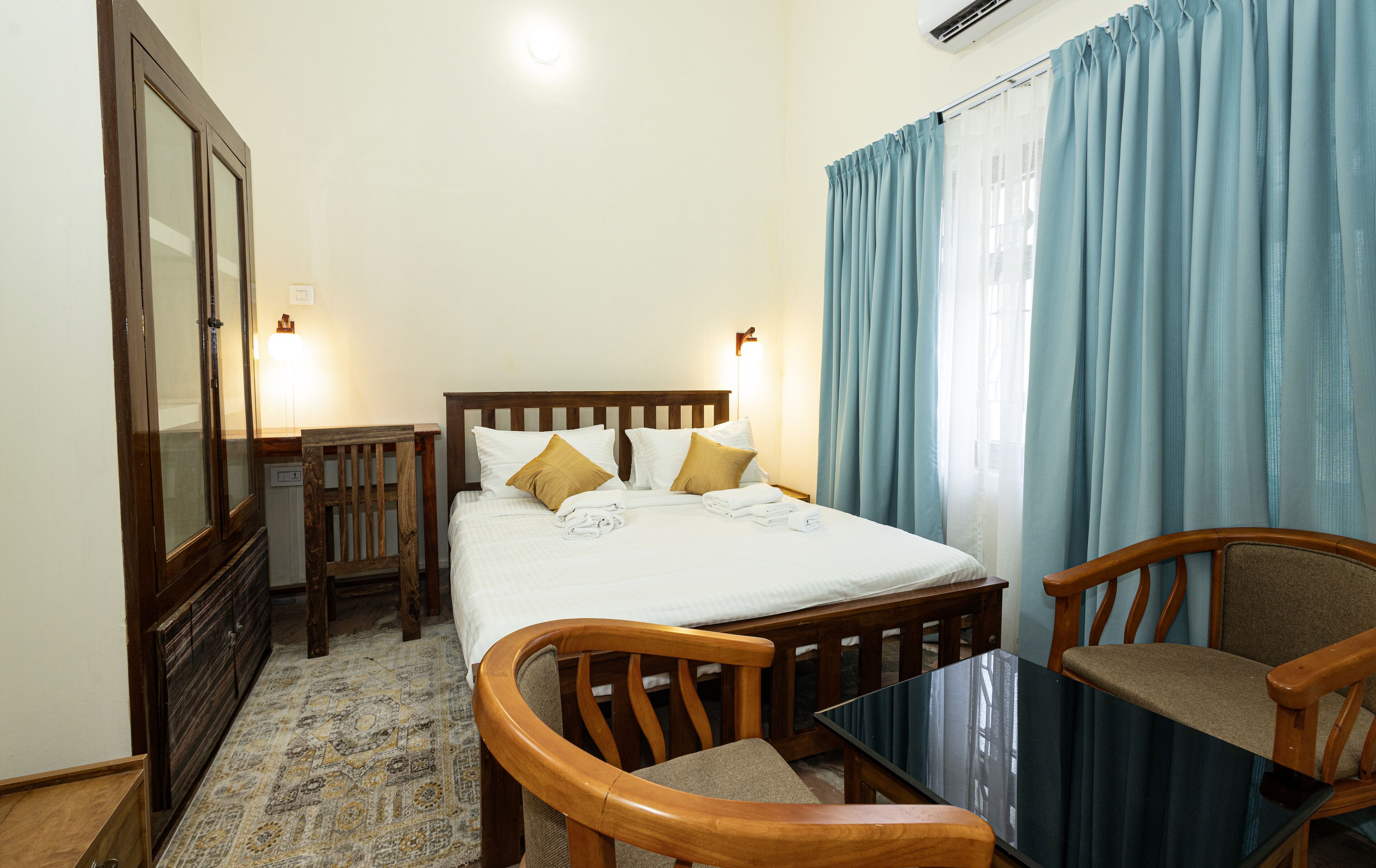 A hotel room with a bed, two chairs, a table, a wardrobe, and a window with curtains.