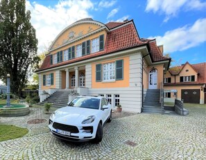 Une maison à deux étages avec un toit recouvert de tuiles rouges, une allée et une voiture blanche garée devant.