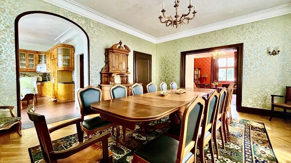 Une salle à manger avec une longue table en bois et des chaises, un lustre, et une vue sur la cuisine.