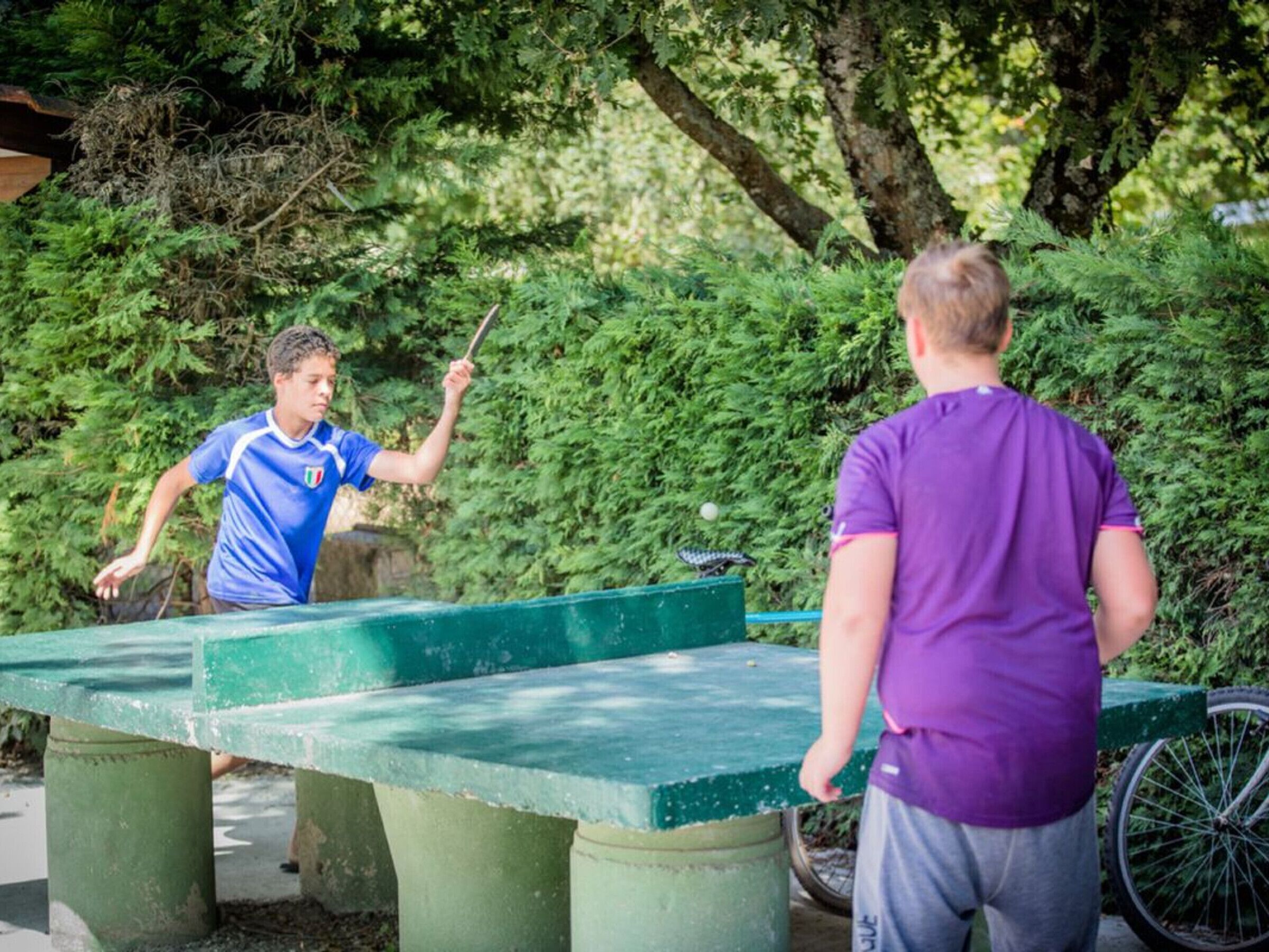 Two people playing table tennis outdoors with a green table and benches, surrounded by greenery.