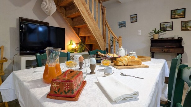 Un comedor con escalera de madera, televisor y una mesa dispuesta con pan, una jarra y una caja decorativa.