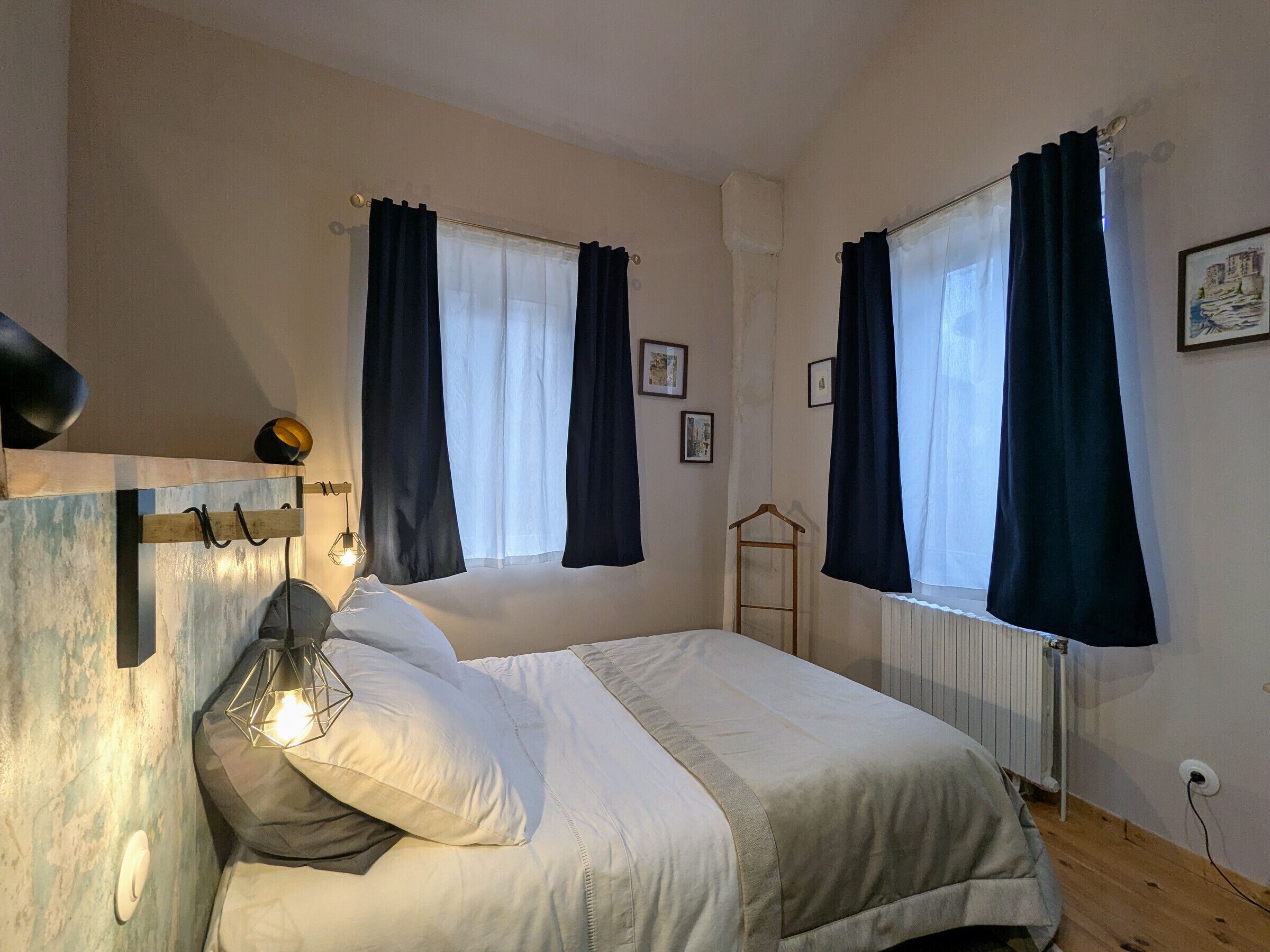 Un dormitorio con cama, lámpara de noche, radiador y dos ventanas con cortinas.