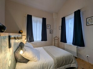 Un dormitorio con cama, lámpara de noche, radiador y dos ventanas con cortinas.