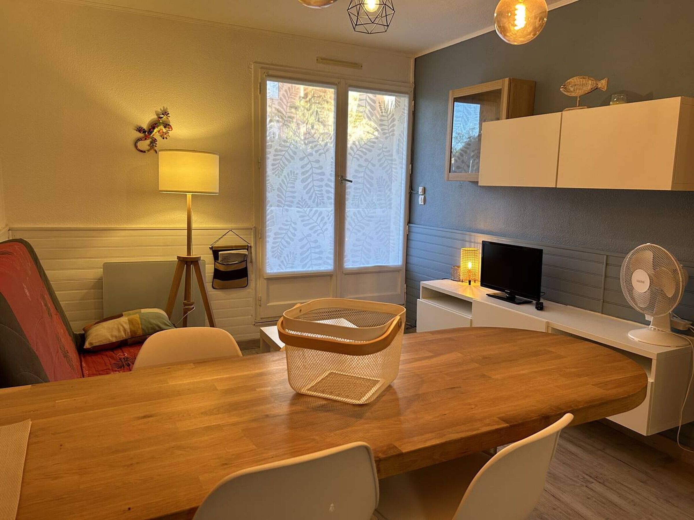 Un coin repas accueillant avec une table en bois, des chaises blanches, un panier, une lampe et un téléviseur.