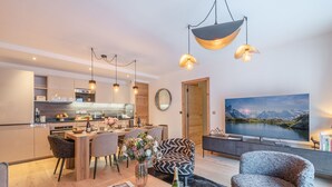 Un espacio de estar moderno con comedor, cocina y un televisor grande que muestra un paisaje pintoresco.