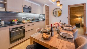 Una cocina moderna con electrodomésticos integrados, un comedor con una mesa de madera preparada para seis comensales, y una sala de estar acogedora con suelo de madera y un espejo en la pared.