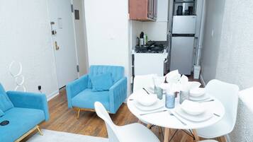 Un salon moderne avec un canapé bleu, une table à manger pour quatre personnes et un coin cuisine équipé d’une cuisinière et d’un réfrigérateur.