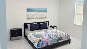 A bedroom with a bed, a ceiling fan, a nightstand, a bedside table, a window with blinds, and a beach-themed painting on the wall. - Enjoyable 1-bedroom house in charming Lehigh Acres/Fort Myers (Lehigh Acres)