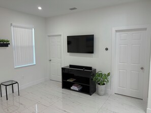 Un salón moderno con un televisor de pantalla plana montado en la pared, un mueble de entretenimiento negro debajo, una planta en maceta y una ventana con persianas.