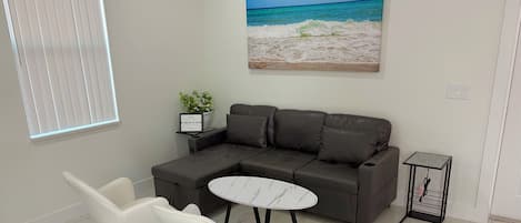 A modern living room with a sectional sofa, two white chairs, a marble coffee table, a painting of a beach, a small plant, and a black side table with a cable holder.
